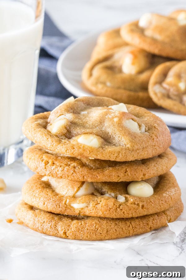 A stack of soft, chewy white chocolate macadamia cookies with golden edges. With a glass of milk and extra plate of cookies in the background.