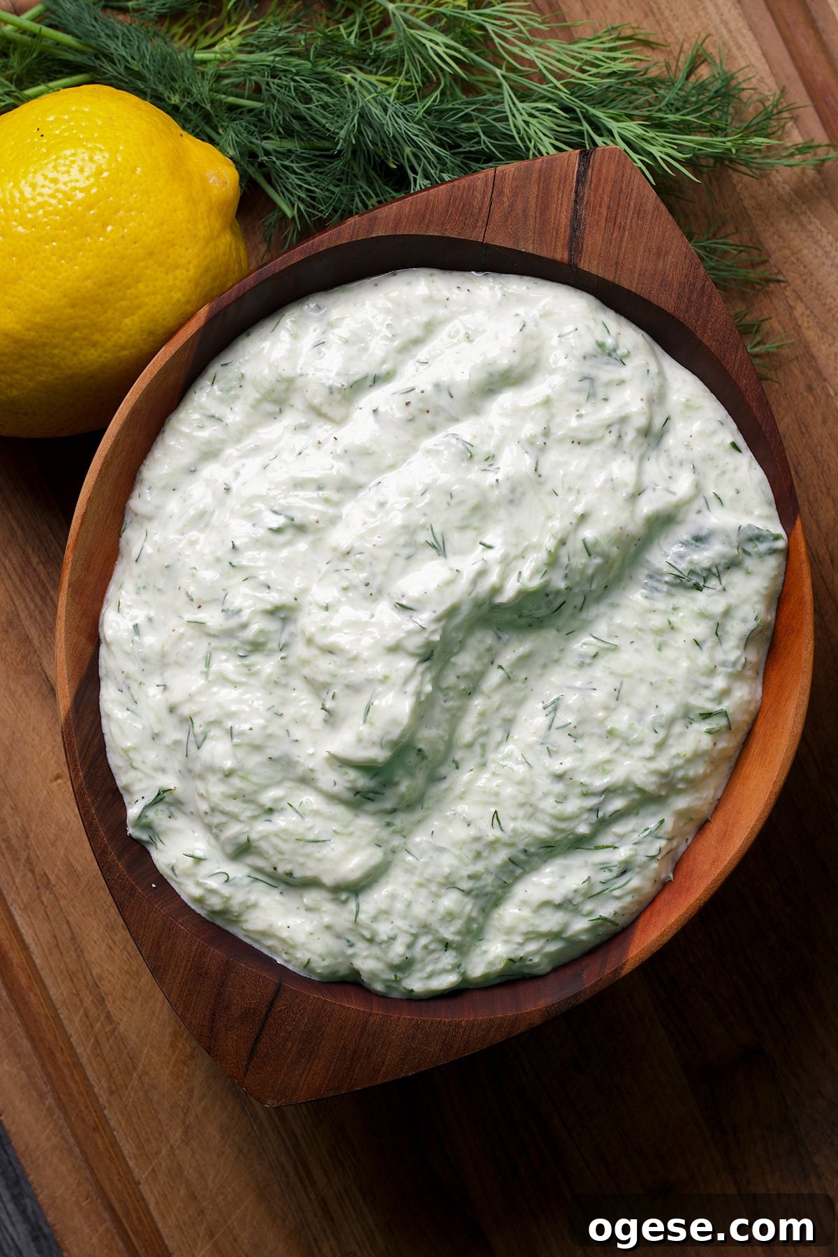 Traditional and Vegan Tzatziki: Two Authentic Recipes You’ll Love 2 A bowl containing tzatziki sauce rests on a wood cutting board surrounded by fresh dill and lemons.