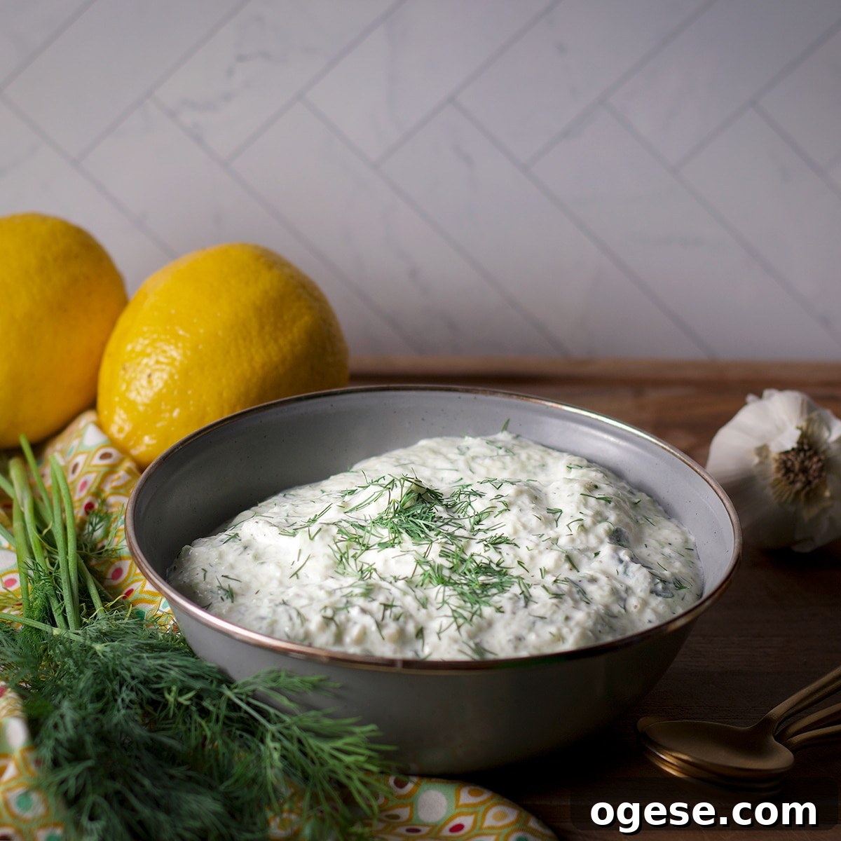 Traditional and Vegan Tzatziki: Two Authentic Recipes You’ll Love 4 A blue bowl containing vegan tzatziki sauce on a wood board surrounded by fresh dill and lemons.