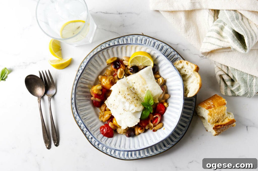 A bowl with a serving of roasted cod with tomatoes and beans next to a slice of bread.