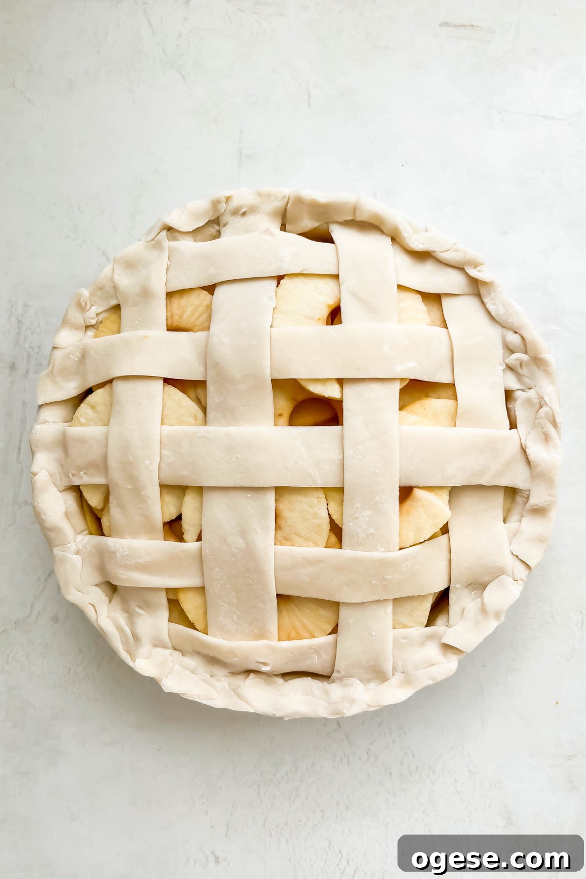 A raw apple pie in a glass pie dish on a white backdrop with a glass of brown butter sugar sauce next to it.