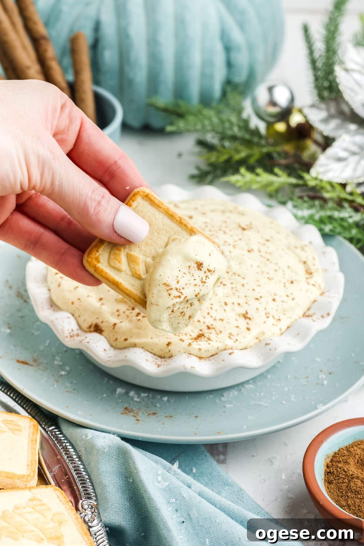 A hand dipping a cookie into a sweet eggnog dip in a bowl on a holiday table.