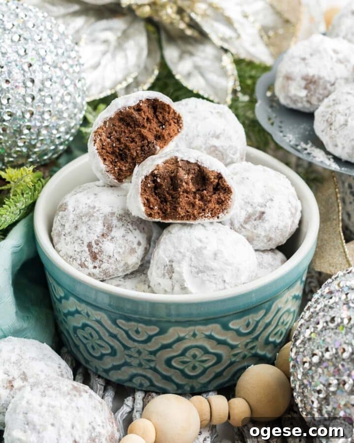 A bowl of chocolate snowball cookies on a table decorated with silver holiday balls and decorations.