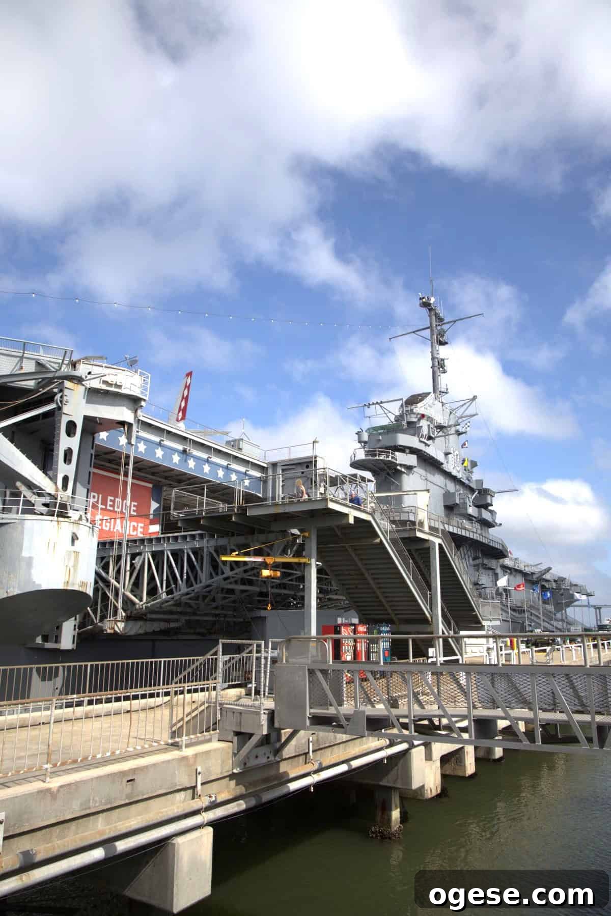 Charleston Travel Guide: Top Attractions, Food & Historic Tours 10 Large naval aircraft carrier docked at port with visible gangways, railings, and a "Pledge of Allegiance" sign; blue sky and clouds in the background—an iconic stop recommended by any Charleston travel guide.