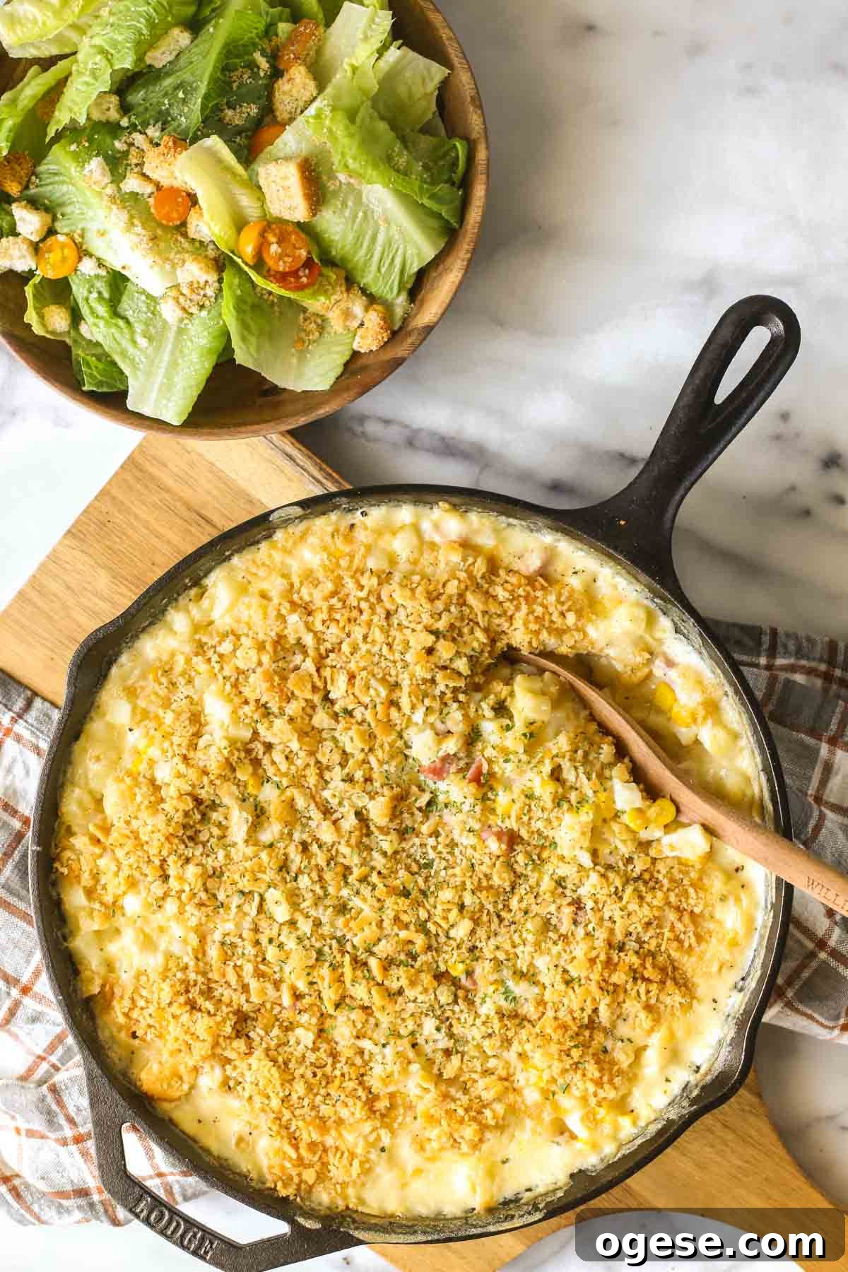 Cheesy Ham & Potato Skillet Recipe for Comforting Weeknight Meals 2 Overhead shot of Cheesy Ham and Potato Skillet in a cast iron skillet sitting on a wood cutting board, with a bowl of salad next to it, all on a marble background.