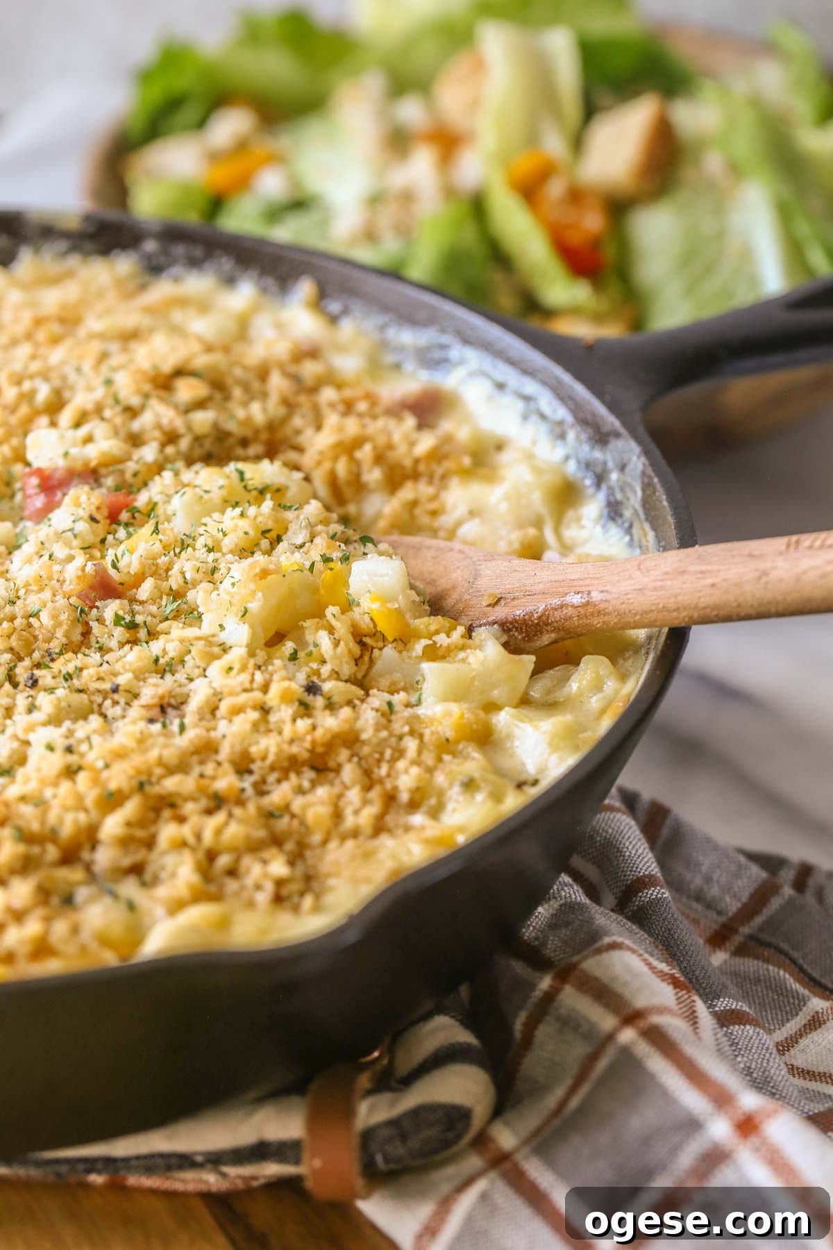 Cheesy Ham & Potato Skillet Recipe for Comforting Weeknight Meals 6 Close up shot of Cheesy Ham and Potato Skillet in a cast iron skillet with a wooden serving spoon in it, and a bowl of salad in the background.