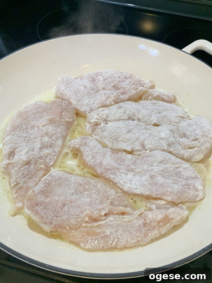 White skillet with raw chicken dredged in flour, cooking in melted butter.