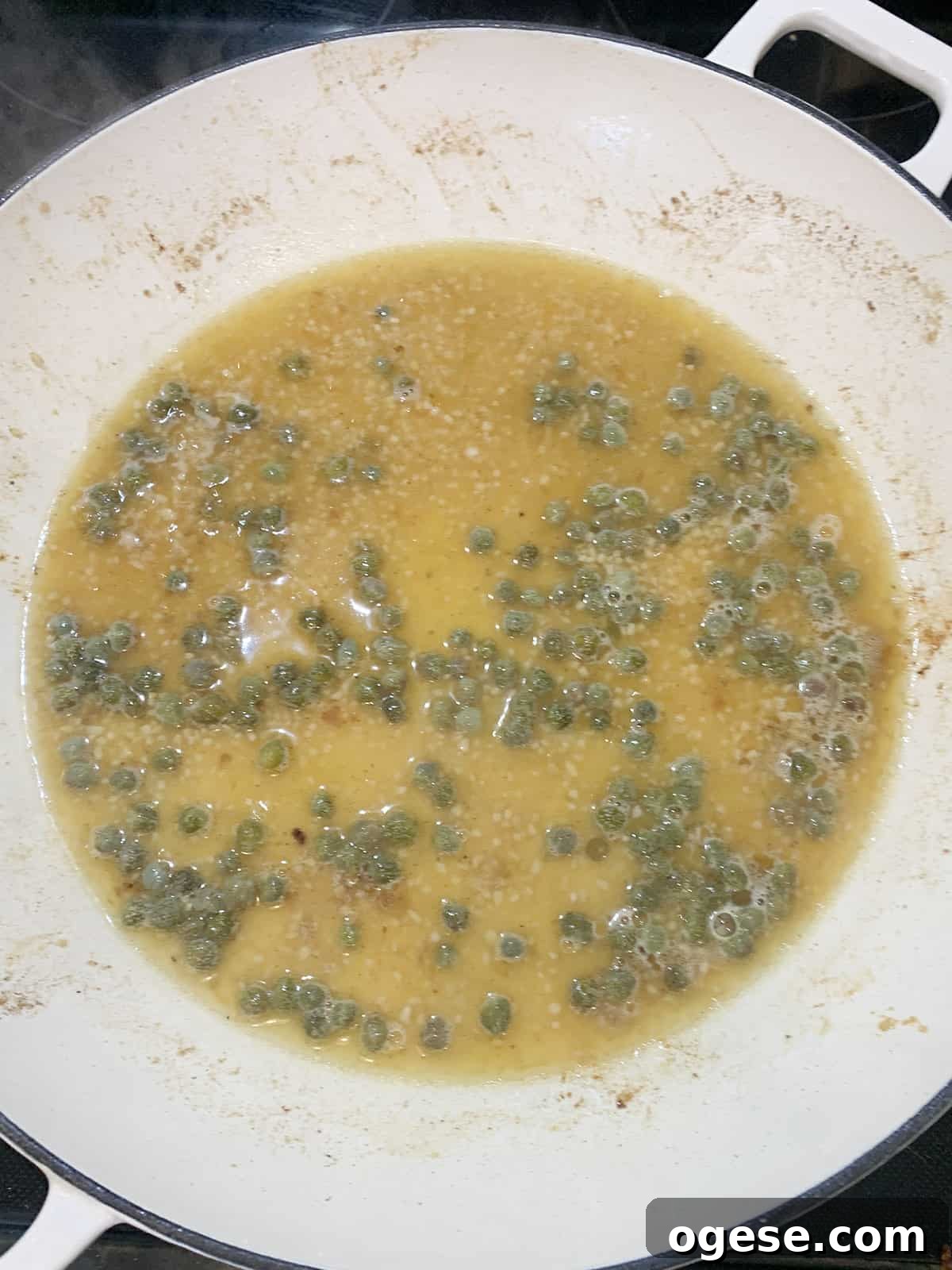 Broth, lemon juice, butter and capers simmering in a white pan on black stovetop.