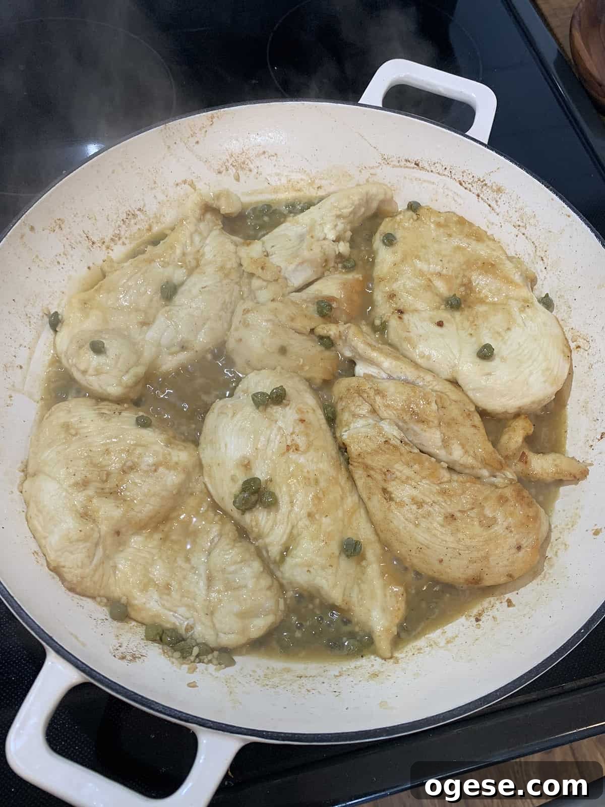 Chicken piccata with sauce and capers cooking in a white pan on black stove.