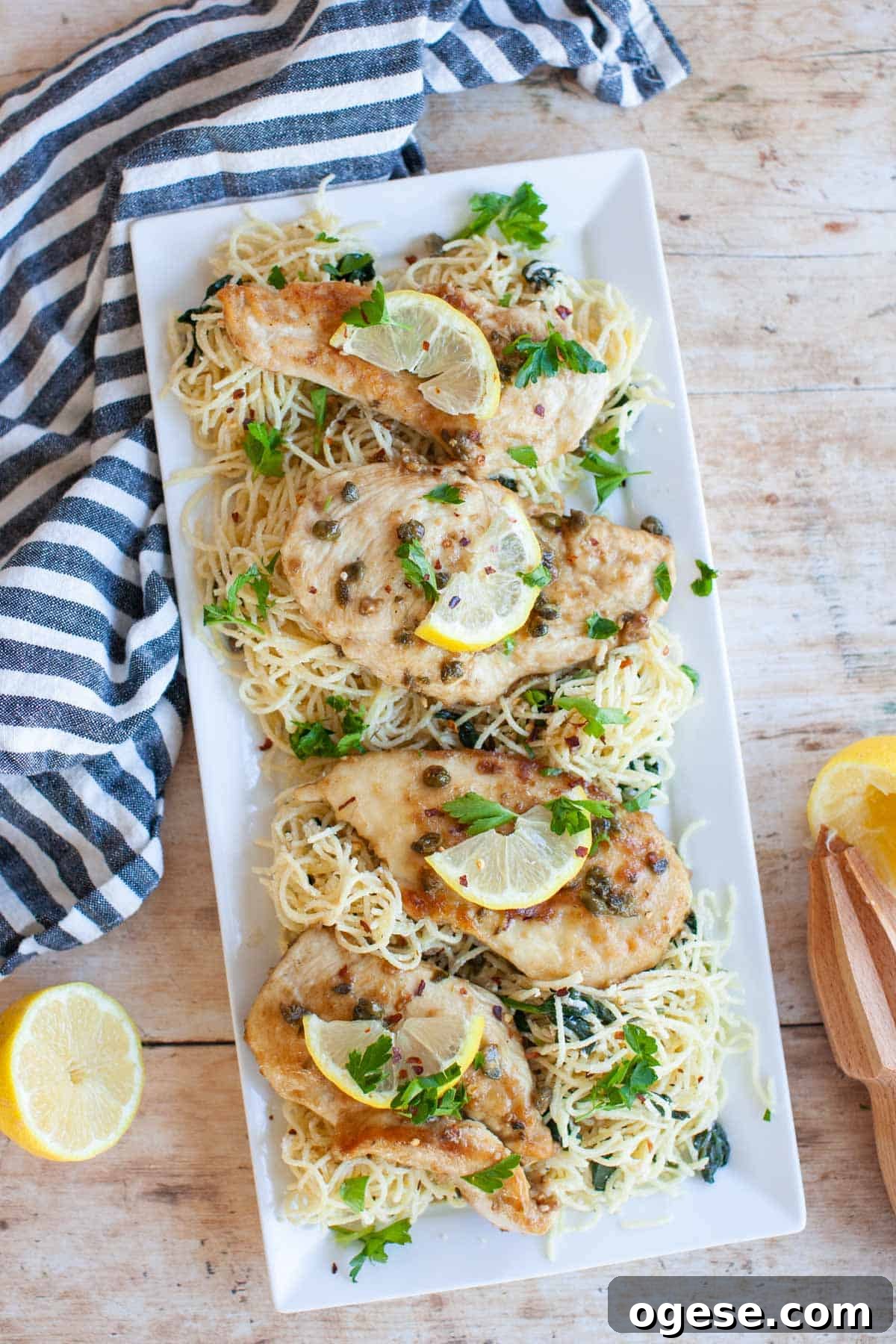 Rectangular white platter with chicken piccata and angel hair pasta, sitting on a wooden board with blue and white napkin, lemon halves and a wooden citrus juicer.
