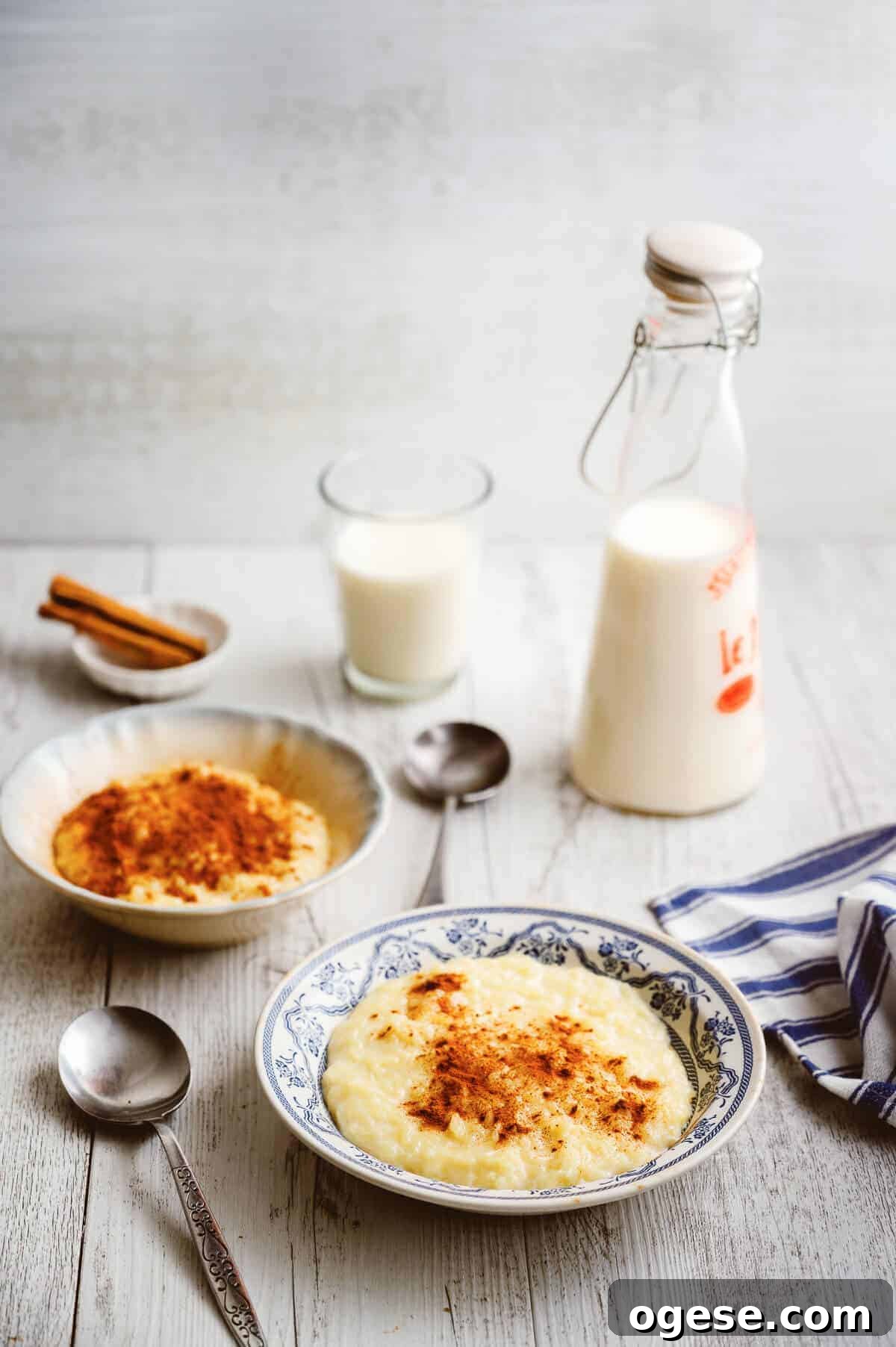 2 bowls of Greek rice pudding on a white table with spoons and pitcher of milk.