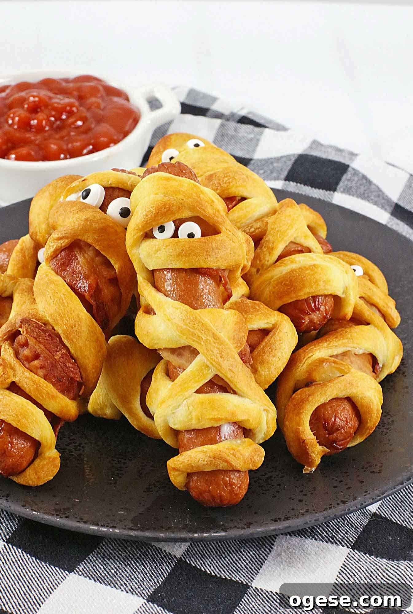 Halloween mummy hotdogs on a black plate with a bowl of ketchup in the background