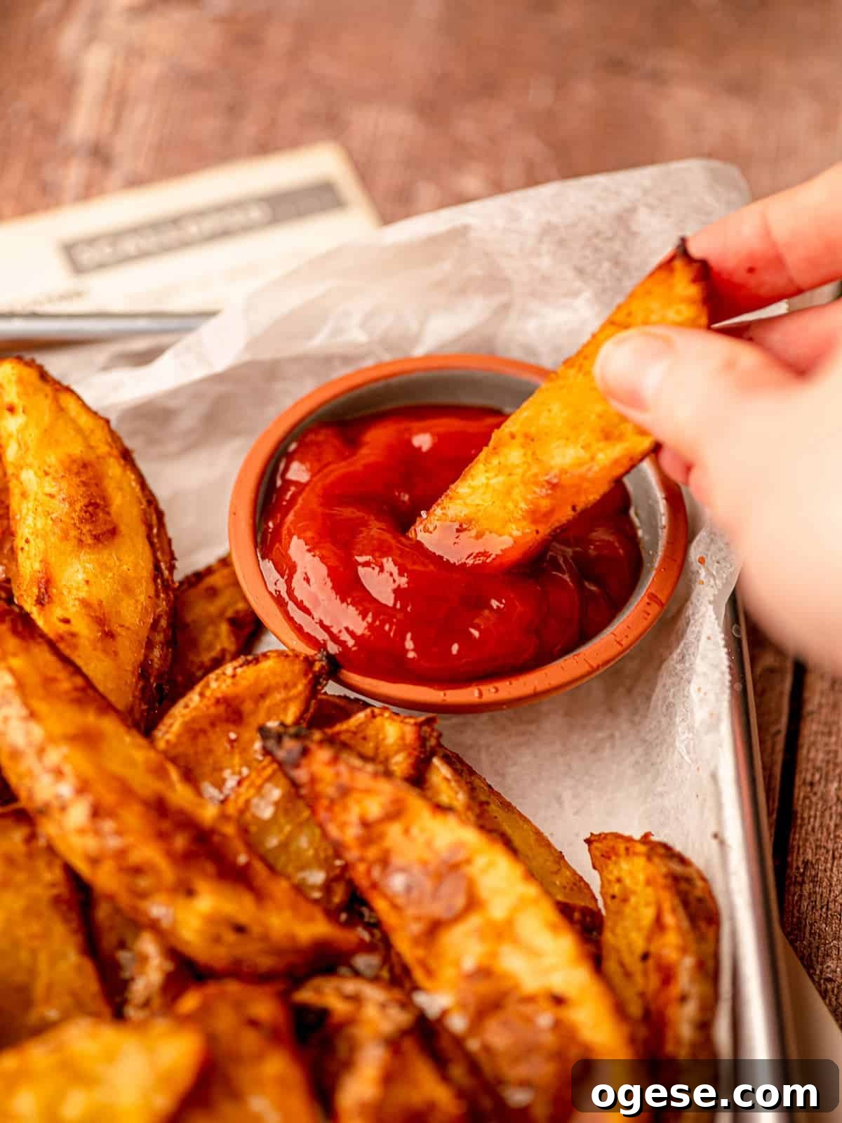 A hand dipping a potato wedge in ketchup.