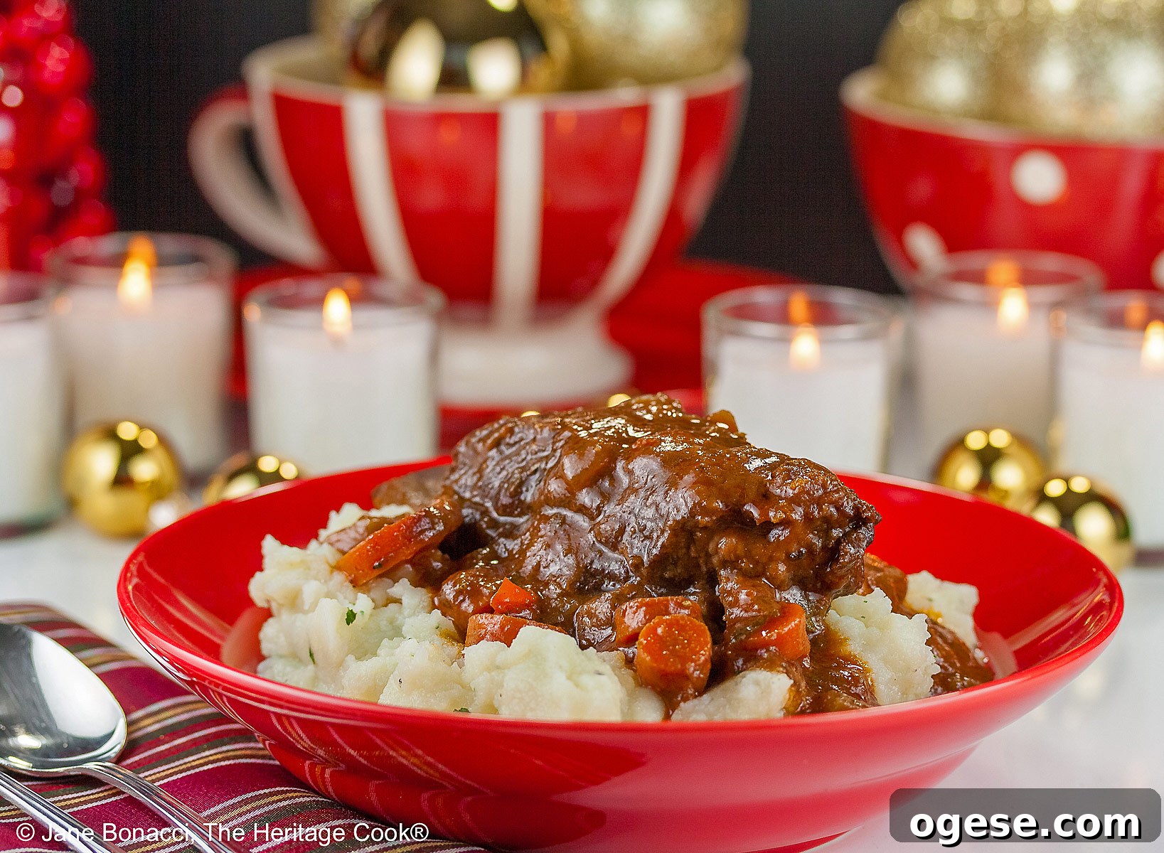 Red Wine-Braised Short Ribs (Gluten-Free) 2 Garlic mashed potatoes lining a red bowl and topped with Red Wine Braised Short Ribs and carrots, in front of festive candles and Christmas decorations with holiday place mats © 2025 Jane Bonacci, The Heritage Cook.