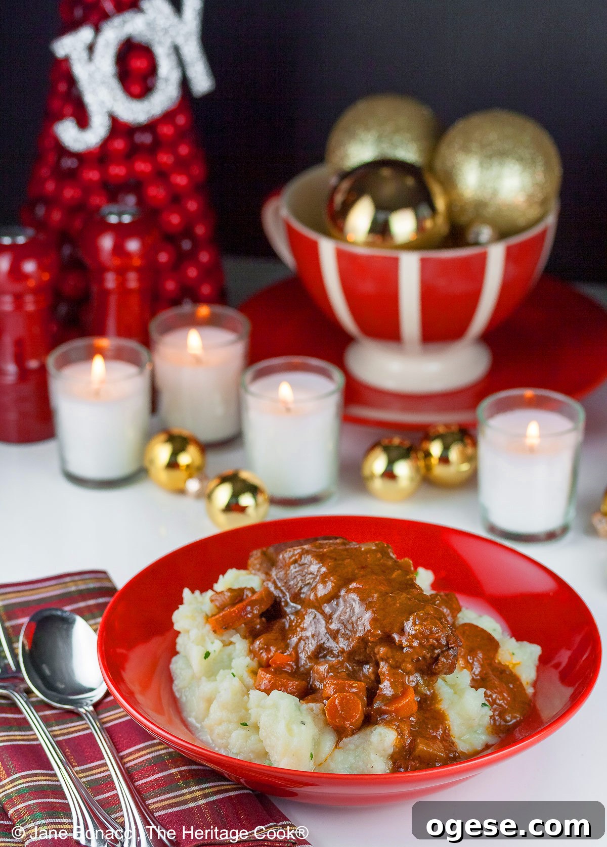 Red Wine-Braised Short Ribs (Gluten-Free) 6 Garlic mashed potatoes lining a red bowl and topped with Red Wine Braised Short Ribs and carrots, in front of festive candles and Christmas decorations with holiday place mats © 2025 Jane Bonacci, The Heritage Cook.