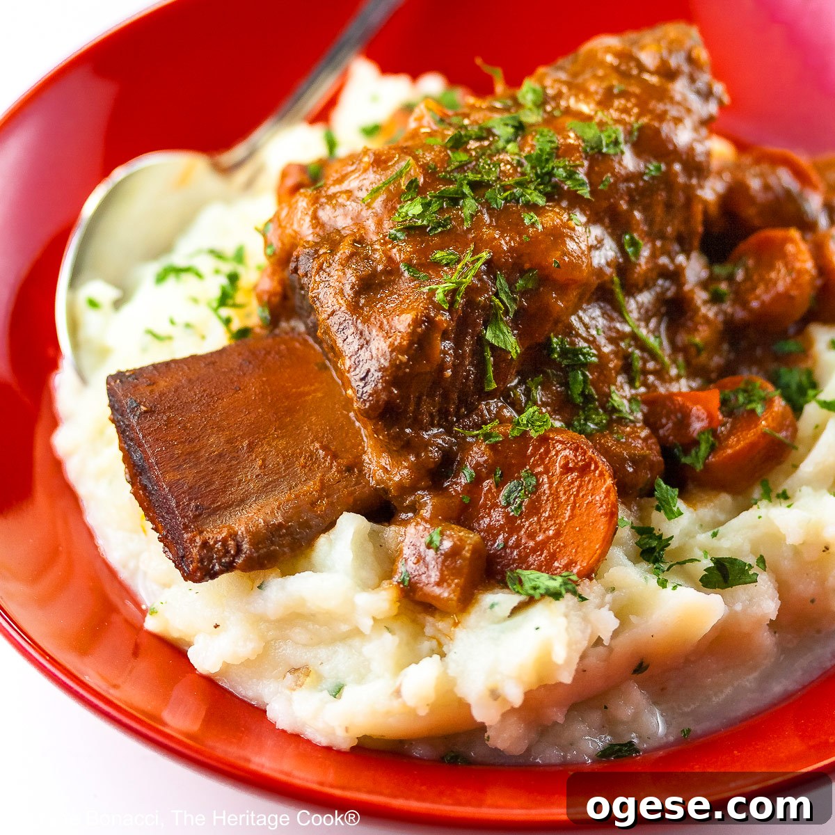 Red Wine-Braised Short Ribs (Gluten-Free) 7 Garlic mashed potatoes lining a red bowl and topped with Red Wine Braised Short Ribs and carrots, in front of festive candles and Christmas decorations with holiday place mats © 2025 Jane Bonacci, The Heritage Cook.