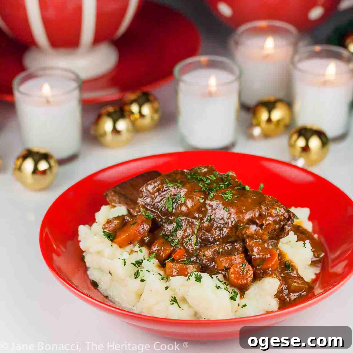Garlic mashed potatoes lining a red bowl and topped with Red Wine Braised Short Ribs and carrots, in front of festive candles and Christmas decorations with holiday place mats © 2025 Jane Bonacci, The Heritage Cook.