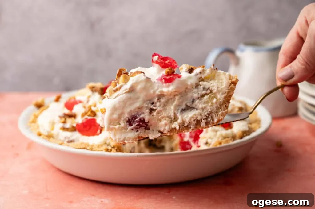 A hand lifts a slice of creamy Million Dollar Pie with cherries and nuts from a pie dish on a pink surface.