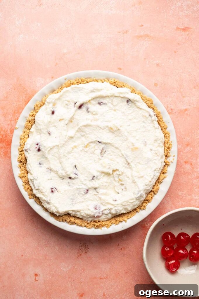 A Million Dollar Pie with a crumb crust and whipped cream topping sits on a pink surface next to a small bowl of maraschino cherries.