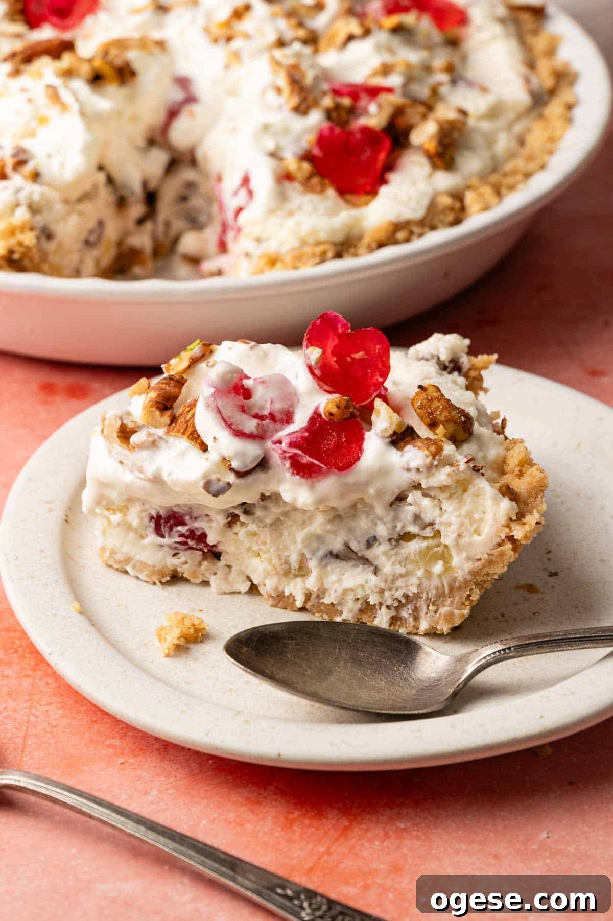 A slice of creamy Million Dollar Pie with chopped nuts and maraschino cherries on a white plate, a spoon beside it, and the remaining pie in the background.