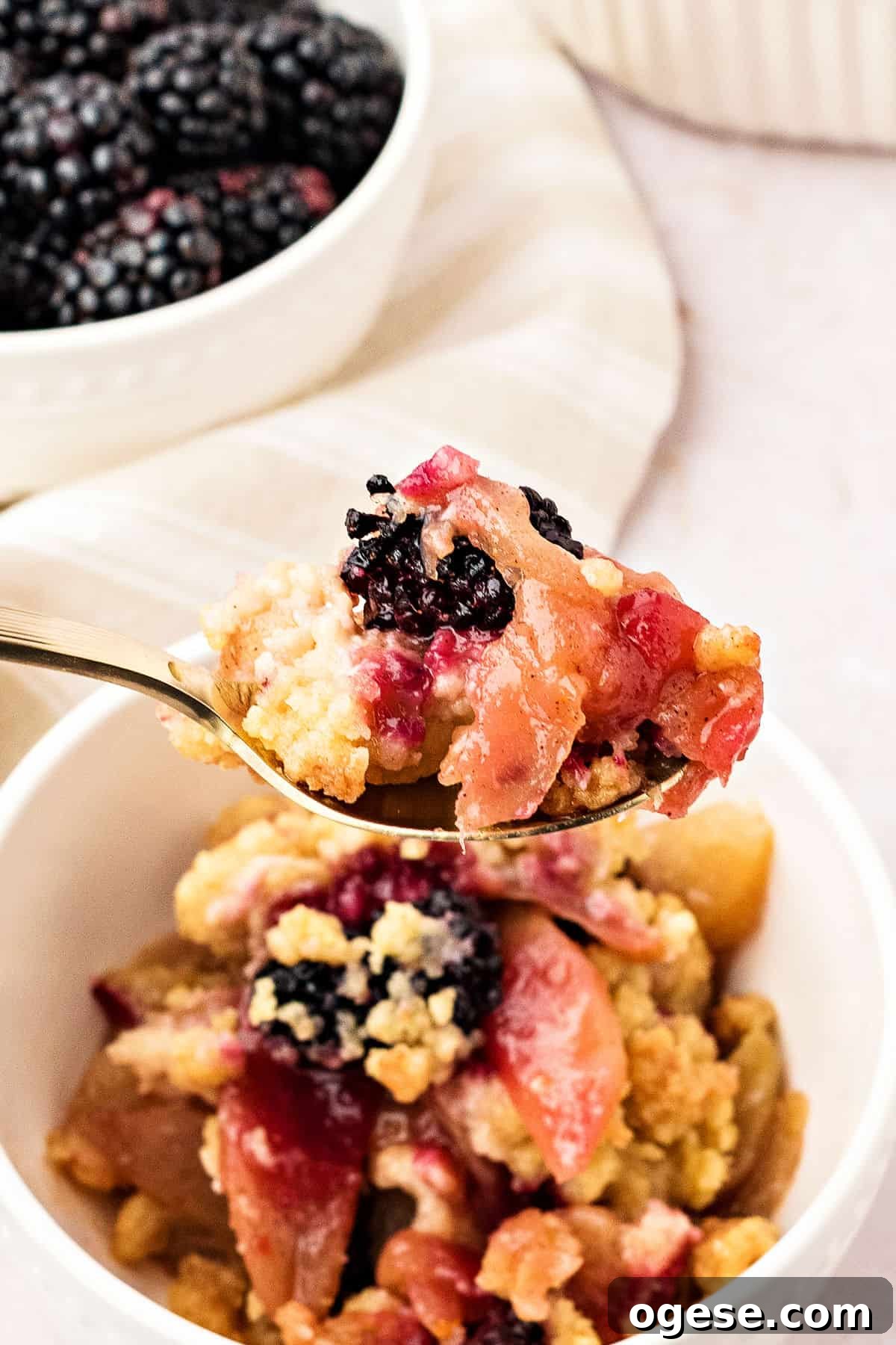 Spoon of apple blackberry crumble over served bowl. Bowl of fresh berries, whitish background.