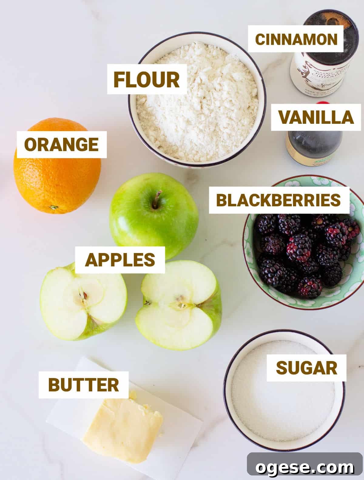 Ingredients for apple blackberry crumble on a white marble surface, including flour, vanilla, orange, butter, sugar.