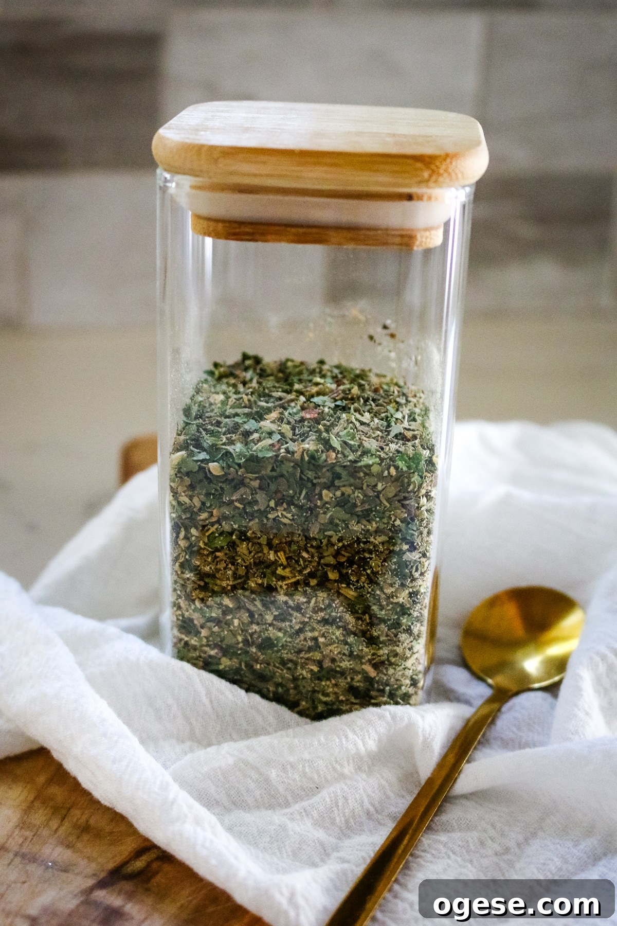 Italian seasoning in a transparent jar, with a spoon next to it.