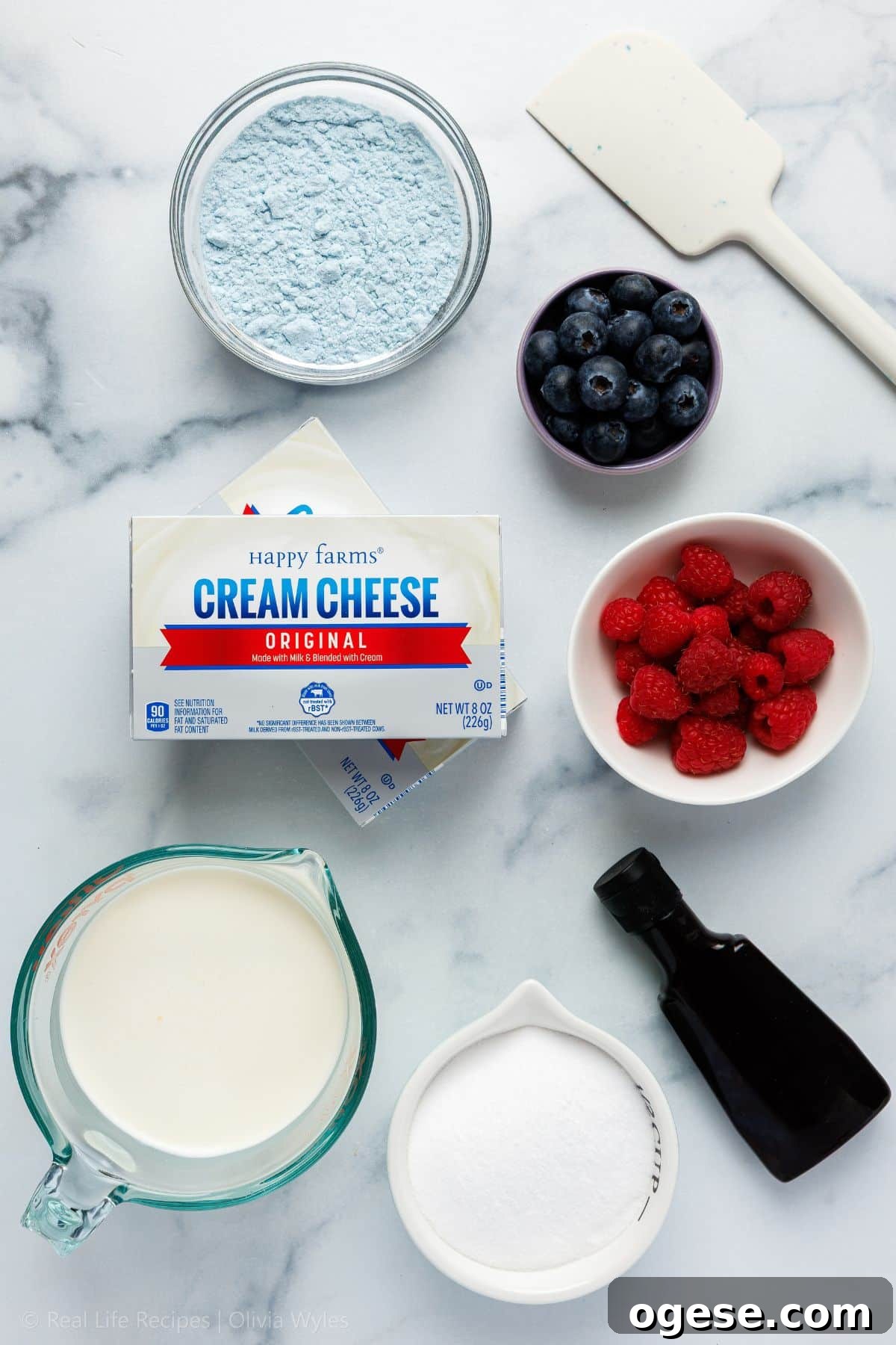Ingredients for no-bake mini blueberry cheesecakes laid out on a marble surface: cream cheese, heavy cream, sweetener, vanilla extract, protein powder, fresh blueberries, and raspberries.