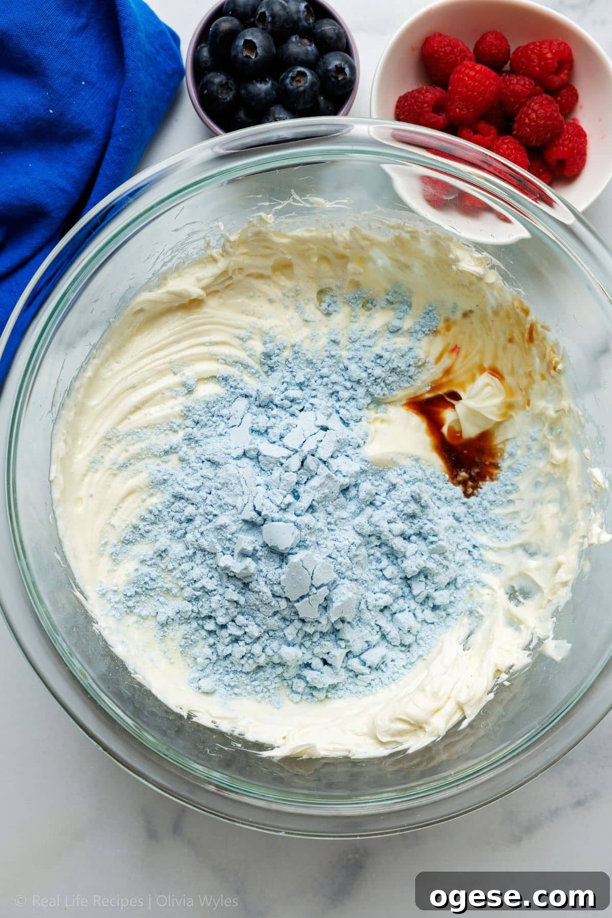 Blue-tinted protein powder and vanilla extract being added to the whipped cream cheese mixture in a glass bowl