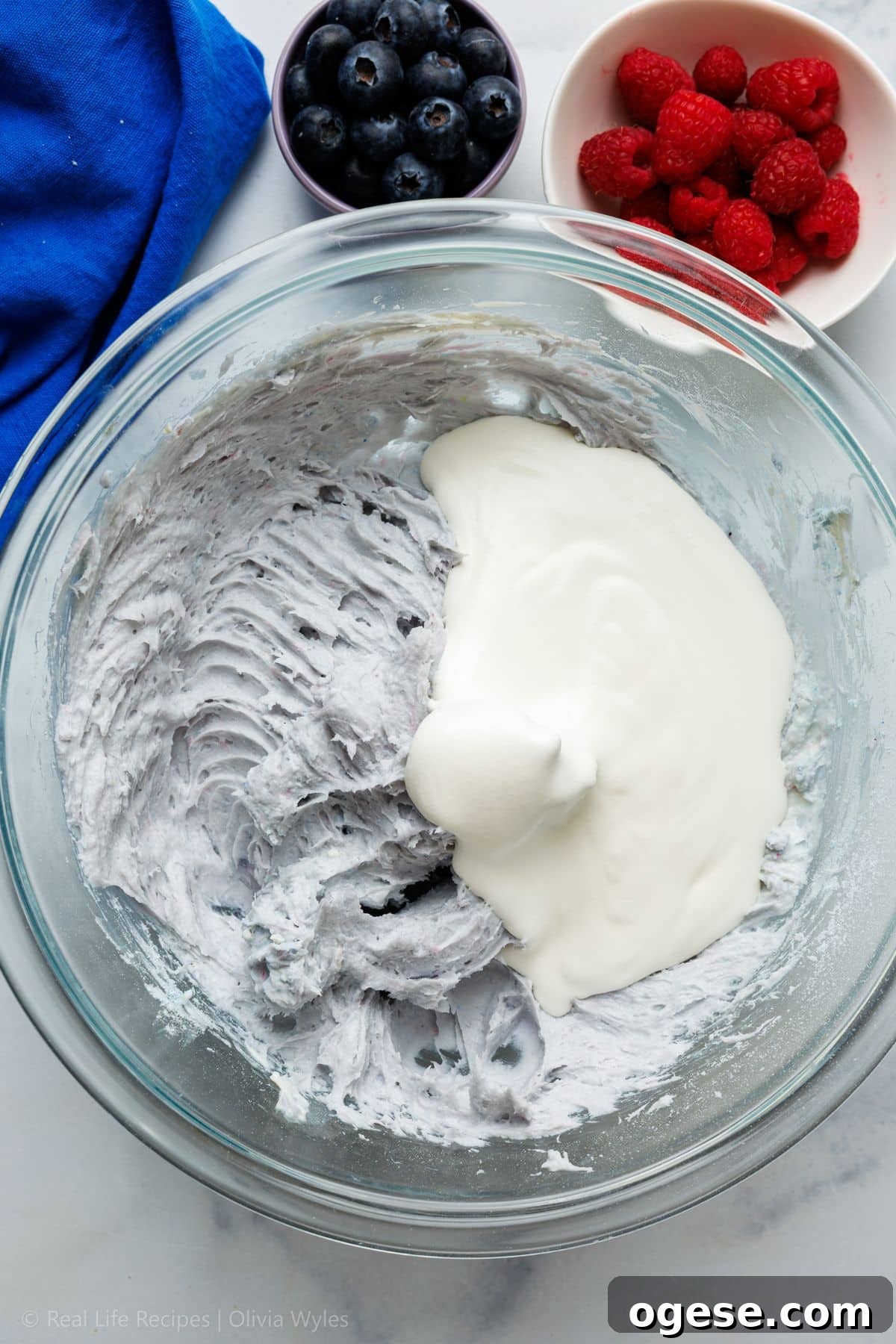 Whipped cream being folded into the cream cheese mixture in a glass bowl, with fresh berries in the background