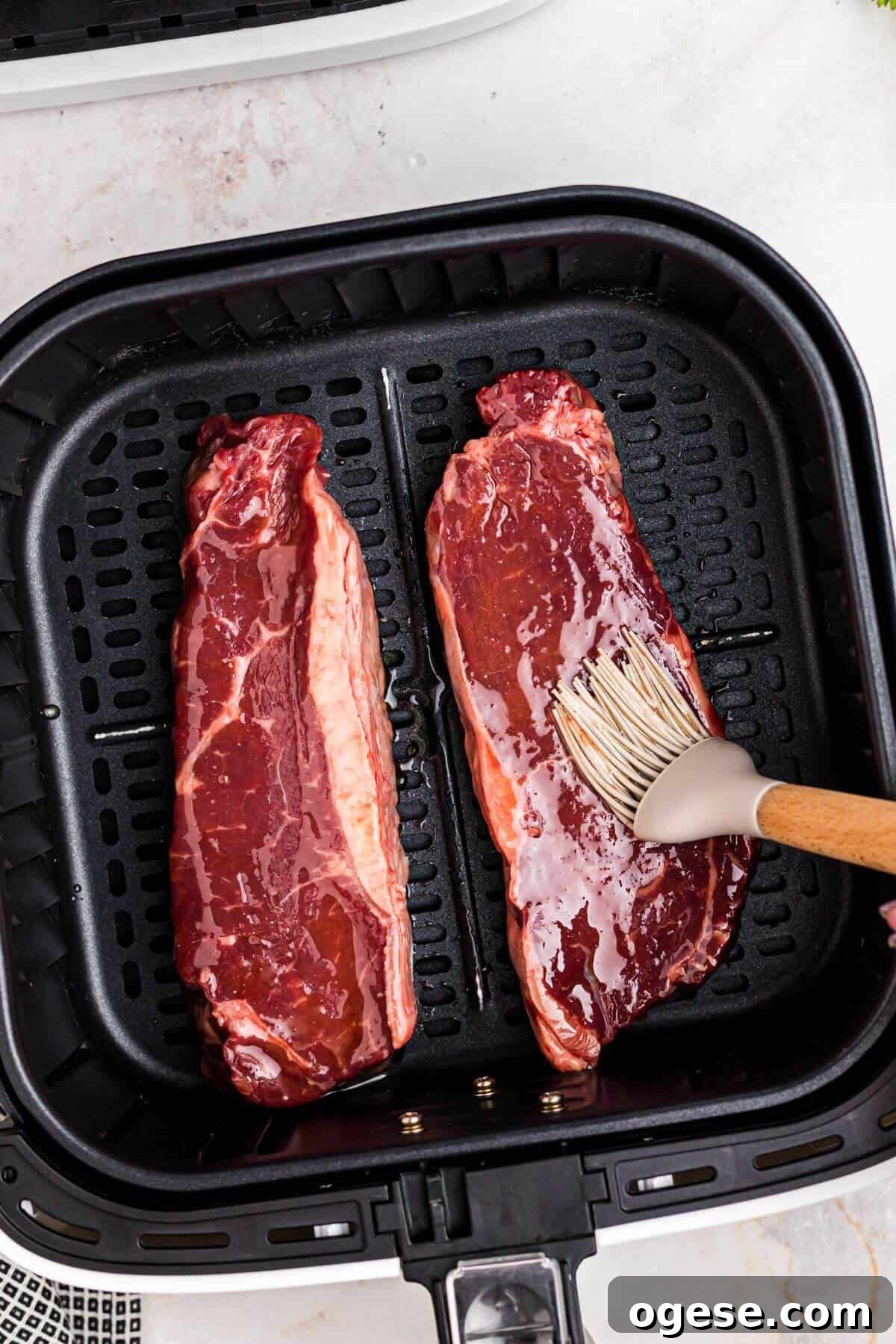 Crispy Air Fryer New York Strip Steak Recipe 5 Uncooked steaks in the air fryer basket being brushed with olive oil.