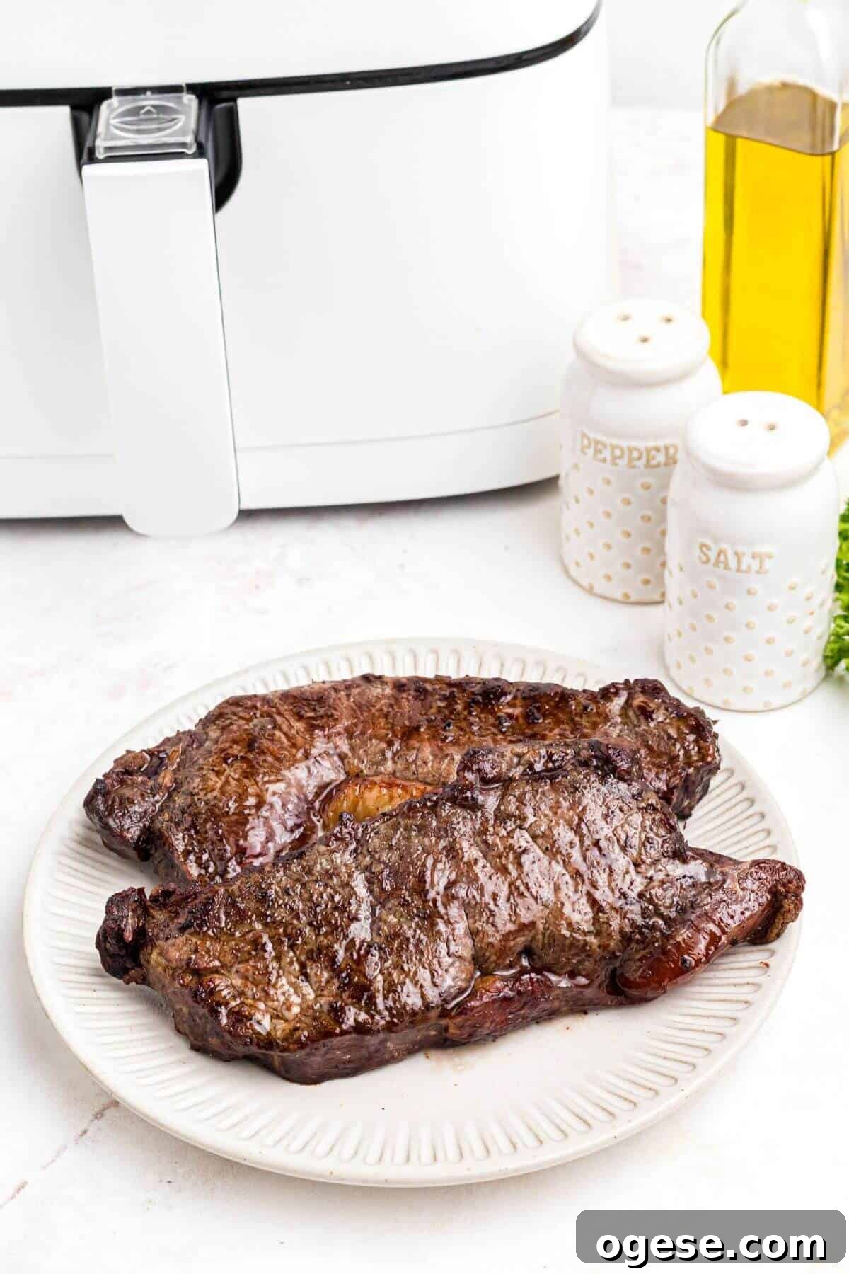 Crispy Air Fryer New York Strip Steak Recipe 8 Juicy cooked air fried strip steaks on a white plate in front of the air fryer.