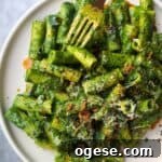 Green pasta sauce with rigatoni on a white plate, with gold fork piercing a piece of pasta.