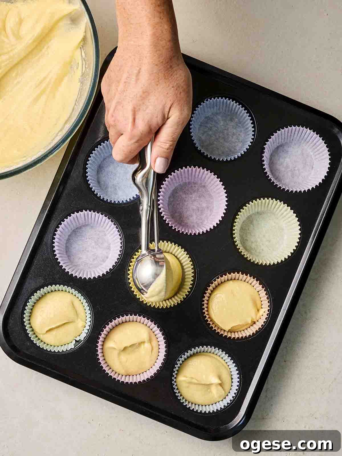 A muffin tray lined with muffin cases, with a hand spooning in cake mixture.