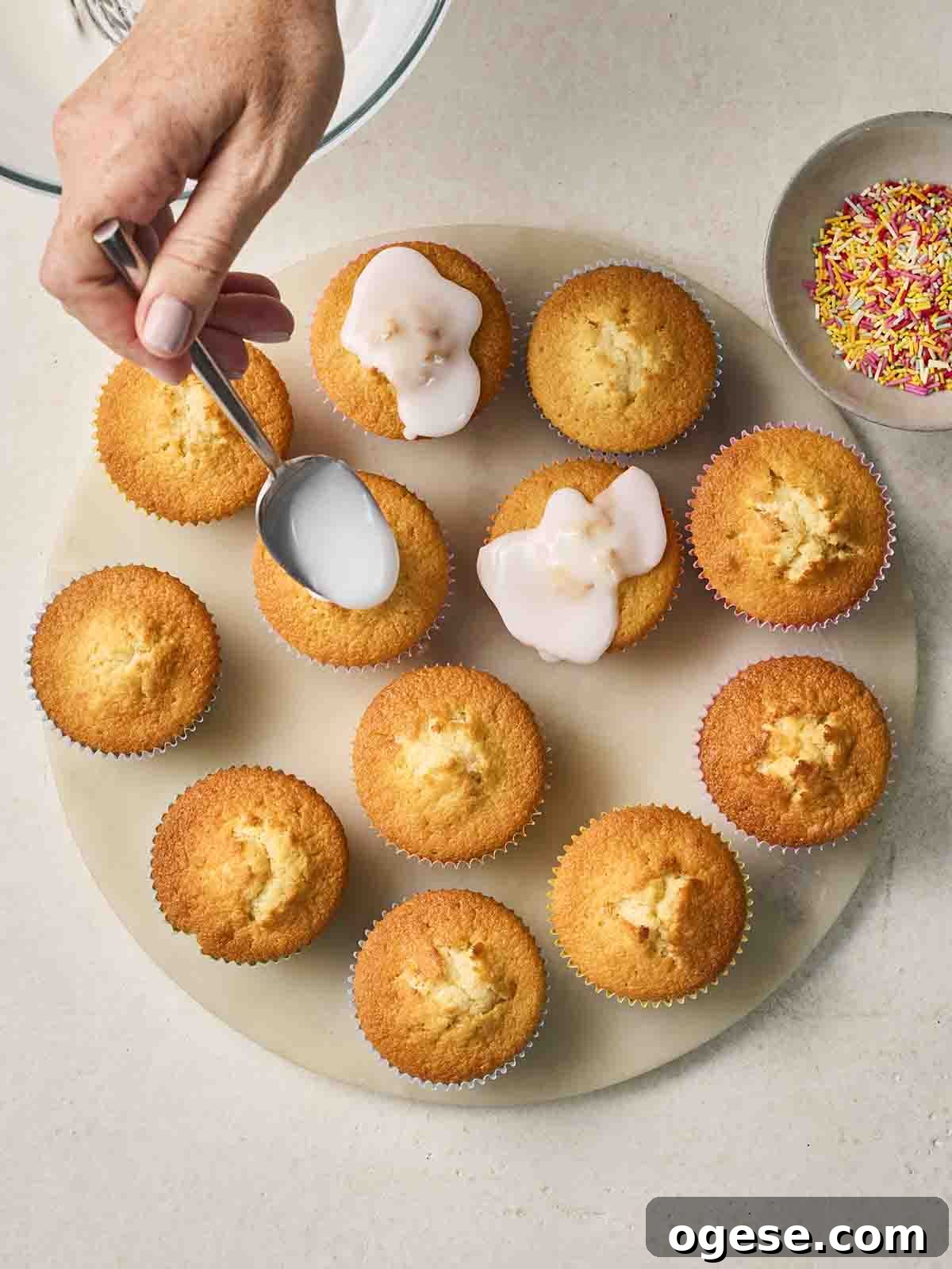 Just-baked Fairy Cakes on a plate with icing being drizzled over the top with a spoon.