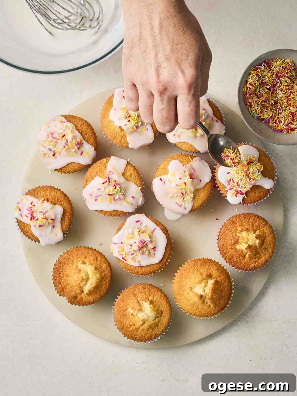 A hand sprinkling hundreds and thousands over the top of a plate of iced fairy cakes.