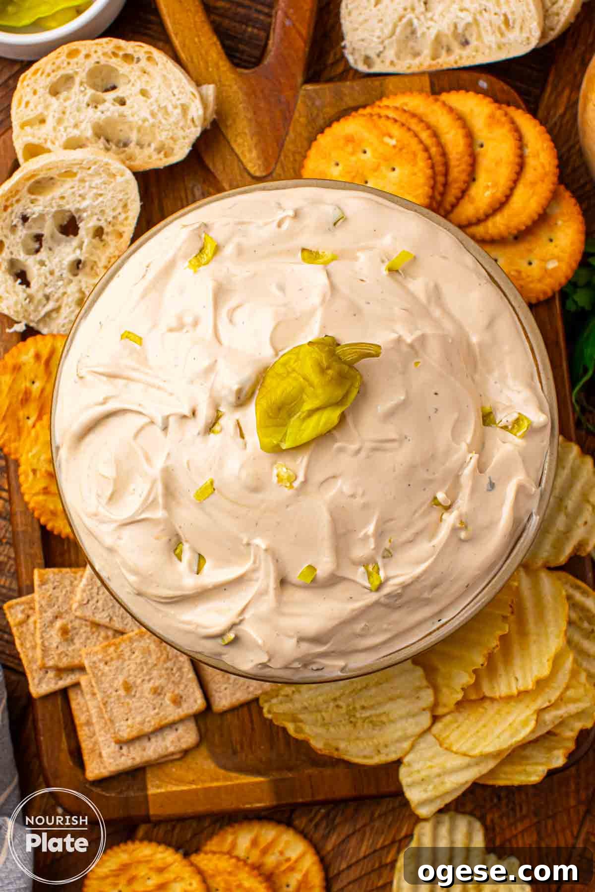 Mississippi Sin Dip in a glass bowl topped with chopped pepperoncini, surrounded by crackers, chips, and sliced bread on a wooden board