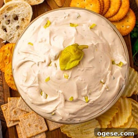Mississippi Sin Dip in a glass bowl topped with chopped pepperoncini, surrounded by crackers, chips, and sliced bread on a wooden board