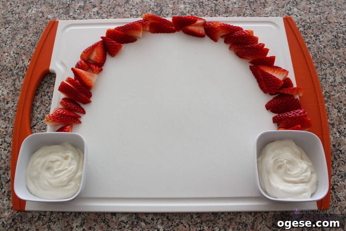 A cutting board on a counter with strawberries arching from one side up to the top and over to the other side, with yogurt dip in small bowls at the bottom of each side of the arch.