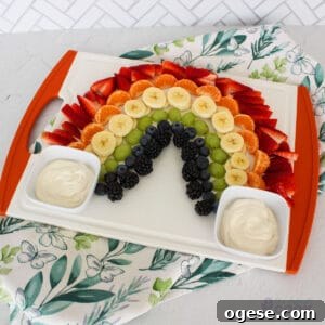 A fruit platter arranged in a rainbow pattern on a white serving tray, with yogurt dips on either side, against a white tile background with a decorative napkin, and 'the default cook' watermark at the bottom right.