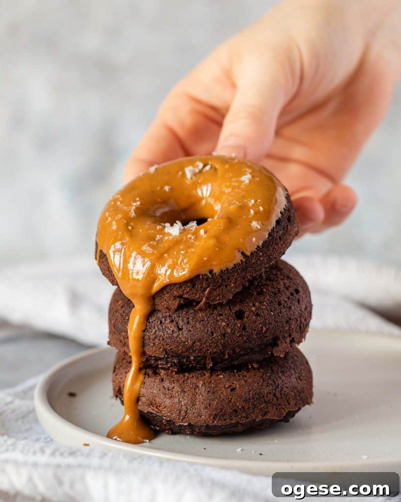 Three Chocolate Baked Protein Donuts stacked on top of each other on a grey plate, there is peanut butter spread on the top on and it's dripping down the side. There is a hand holding the top one.