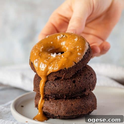 Three Chocolate Baked Protein Donuts stacked on top of each other on a grey plate, there is peanut butter spread on the top on and it's dripping down the side. There is a hand holding the top one.