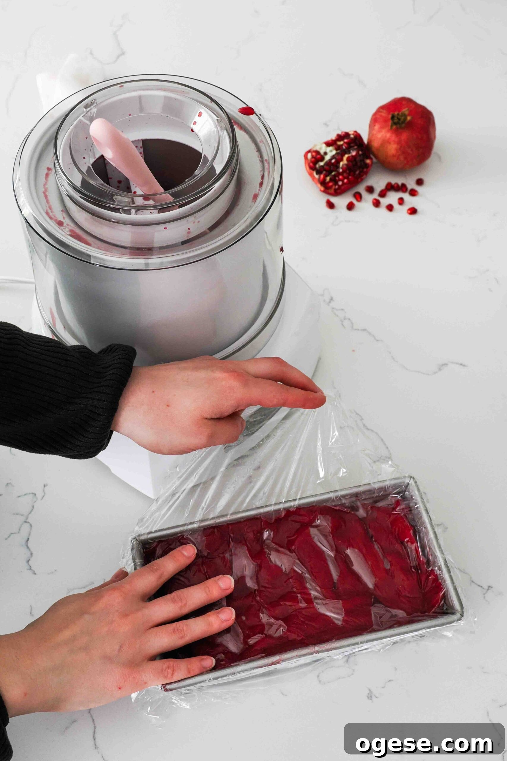 Two hands place plastic wrap over a loaf tin filled with pomegranate sorbet.
