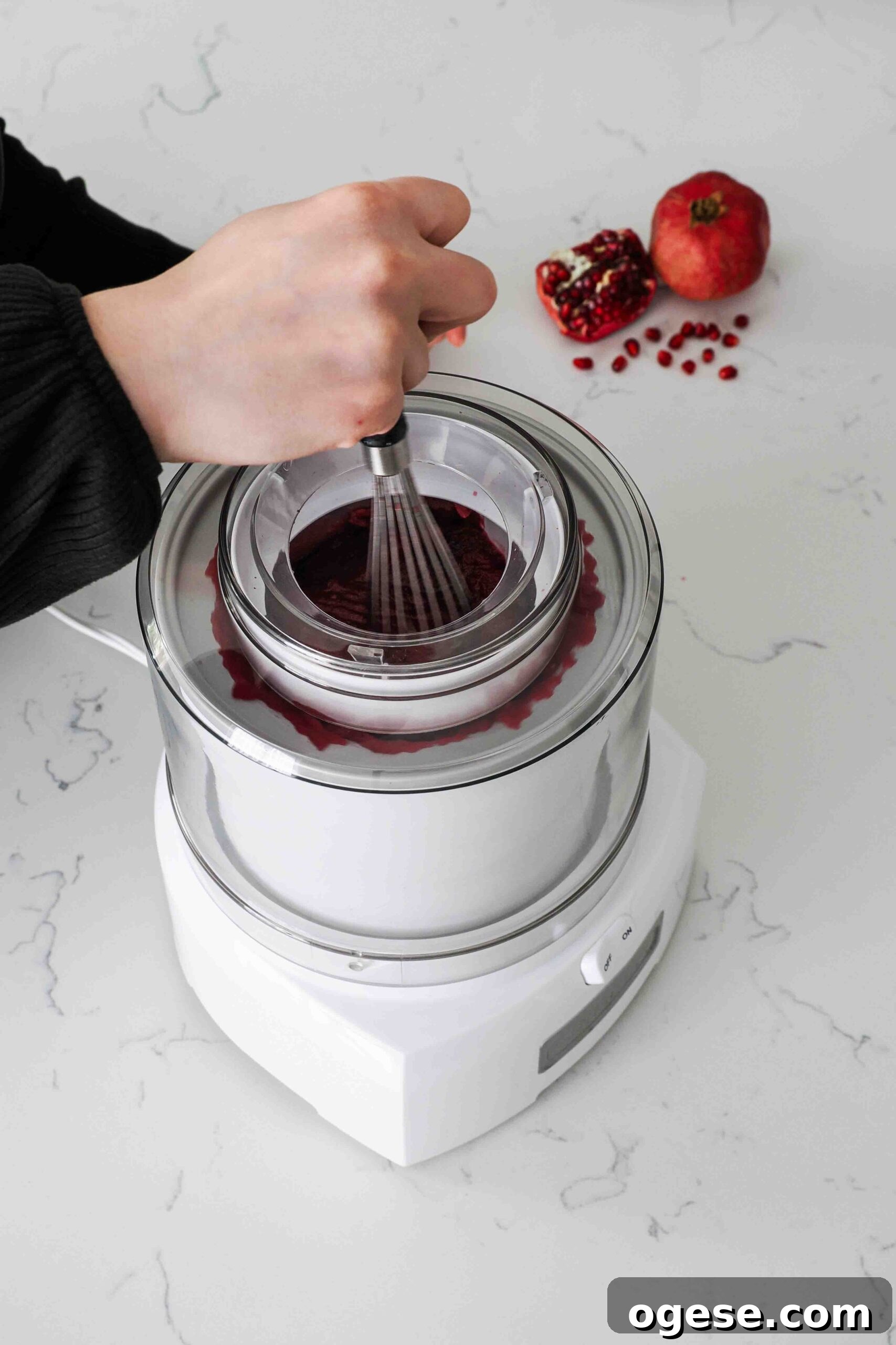 A hand uses a whisk to break up frozen chunks of pomegranate sorbet in an ice cream maker.