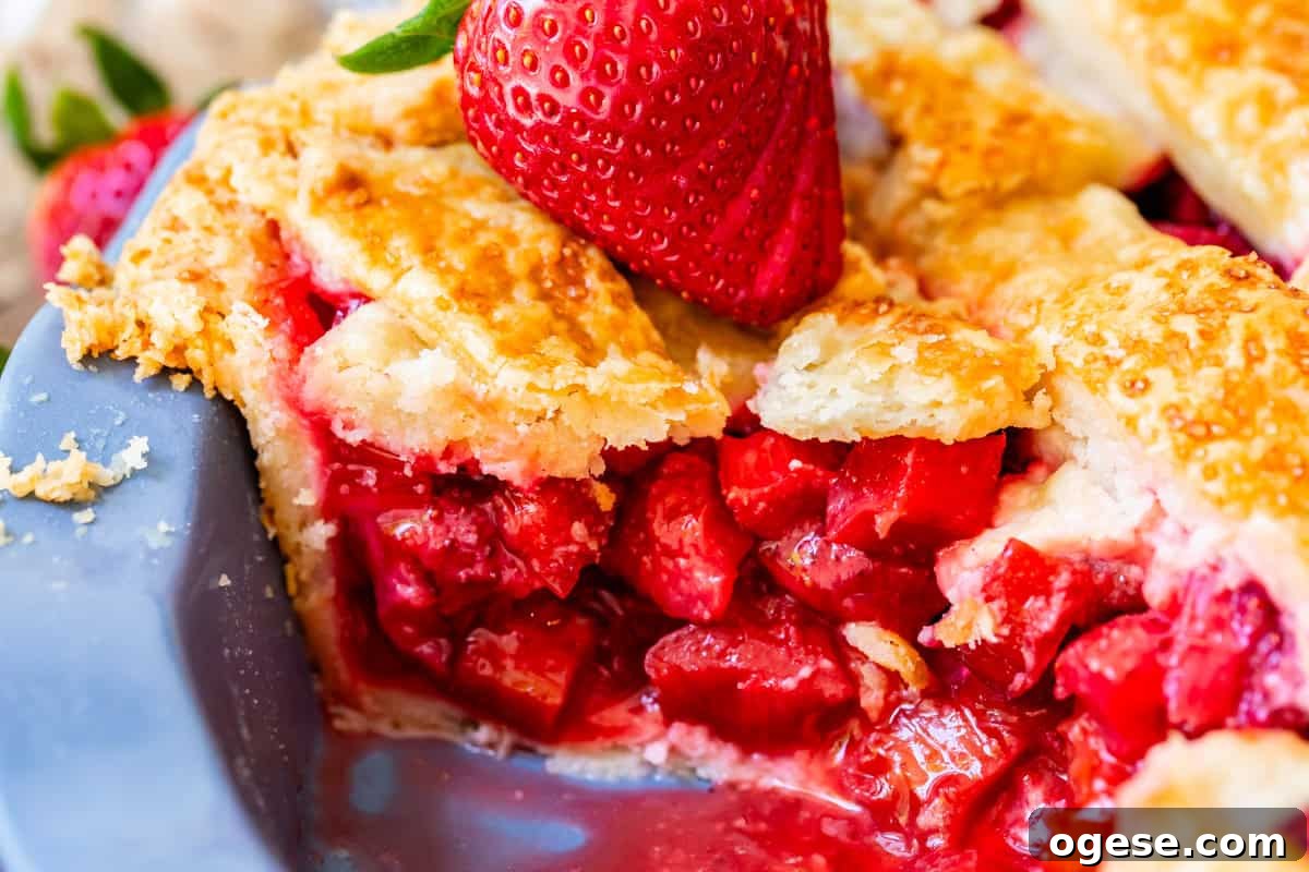 close up of side view of a slice of strawberry rhubarb pie with flaky crust and strawberry garnish.