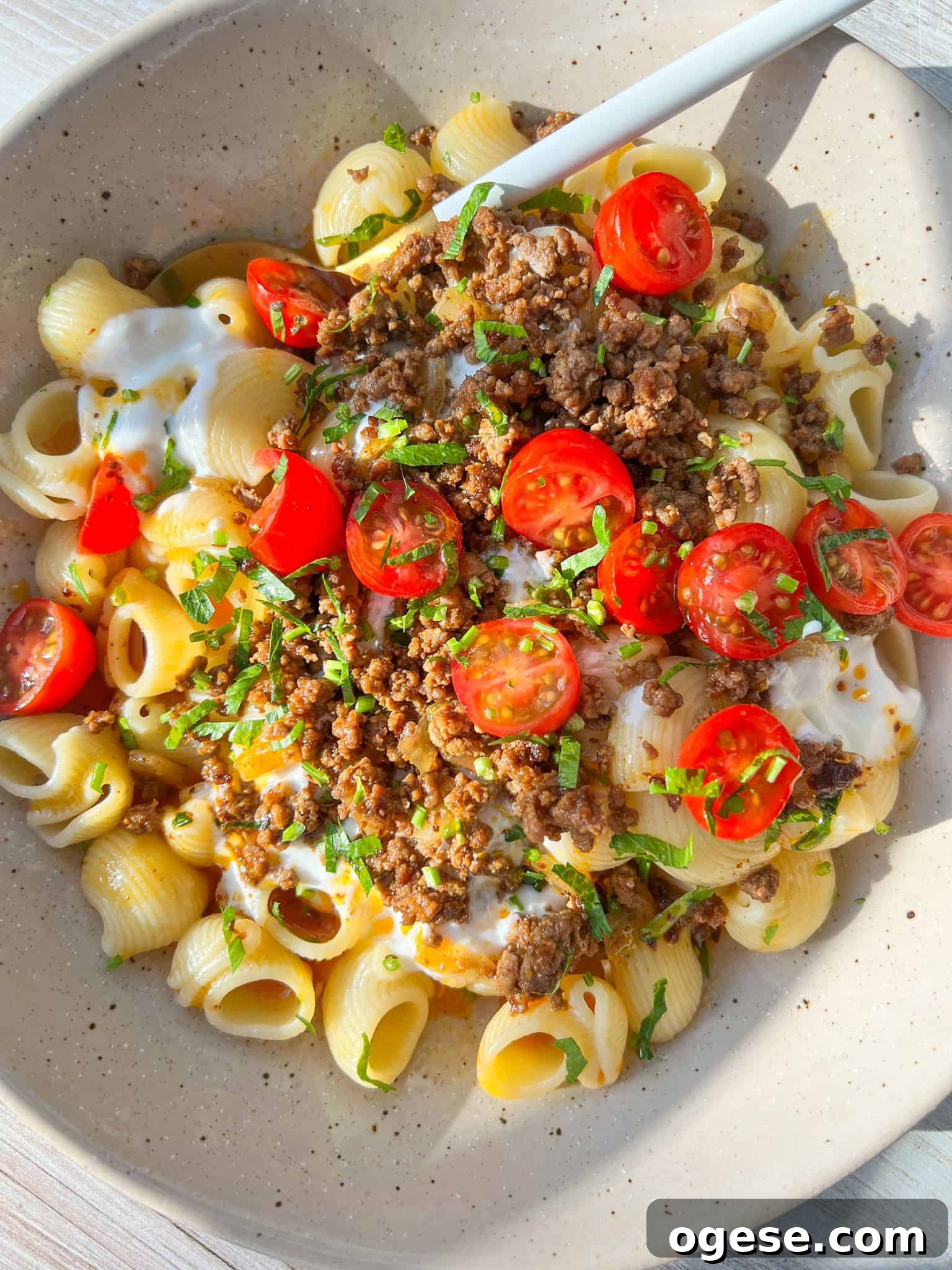 A bowl of turkish pasta topped with seasoned ground beef, sliced cherry tomatoes, dollops of sour cream, and chopped fresh herbs. The dish is served with a spoon on a light-colored plate.
