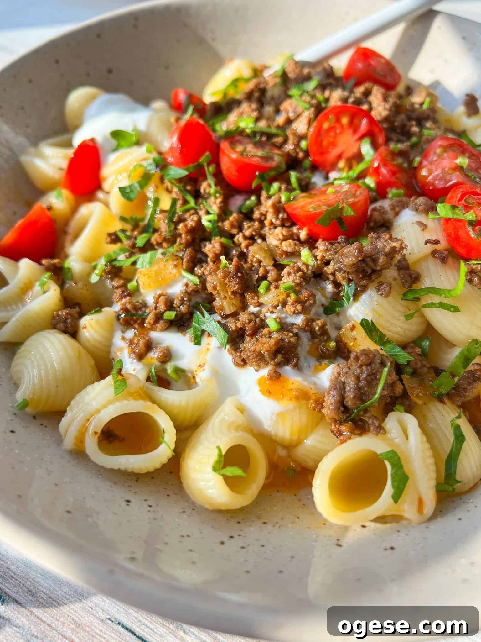 A bowl of Turkish pasta with creamy sauce, topped with seasoned ground meat, cherry tomato halves, and fresh parsley. The dish is served in a light-colored bowl with a spoon. Sunlight casts a warm glow over the meal.