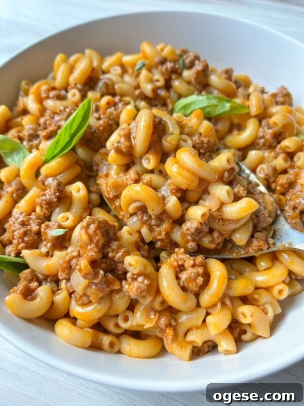 A bowl of macaroni pasta with ground beef in a creamy tomato sauce, reminiscent of homemade Hamburger Helper, garnished with fresh basil leaves. A spoon rests in the bowl, ready to eat.