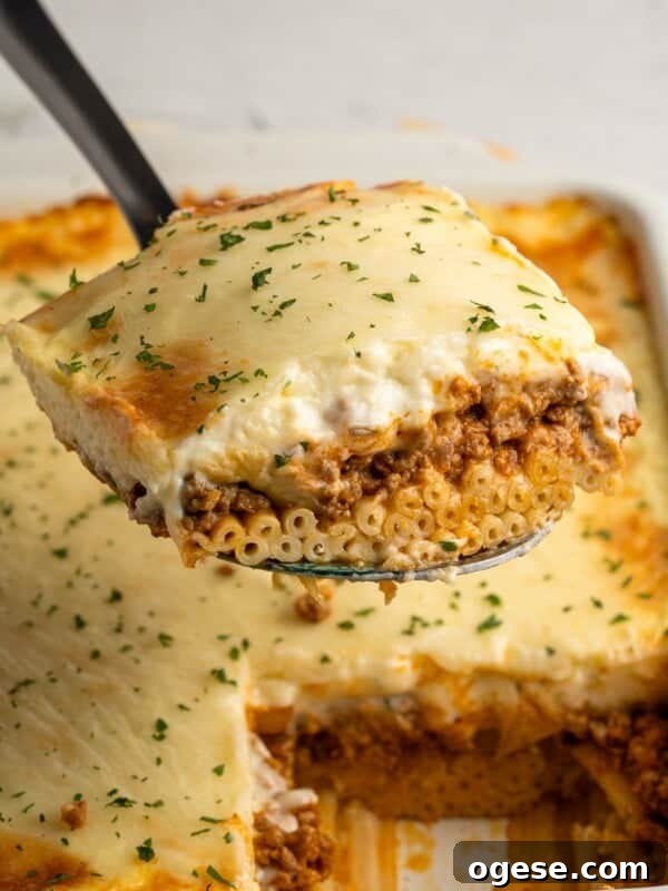 A close-up of a slice of pasta topped with melted cheese and garnish, being lifted from a baking dish. The pasta is layered with a meat sauce and creamy topping, with parsley sprinkled on top. The dish is set against a neutral background.