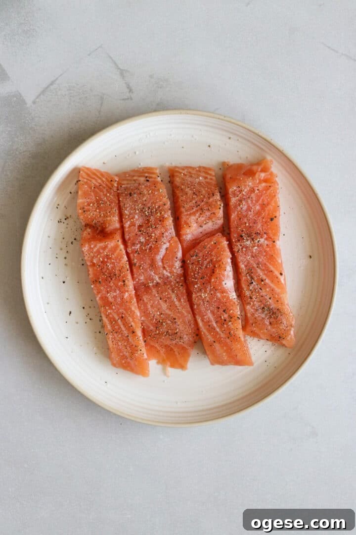One-Pan Lemon Salmon and Orzo Recipe 5 Four salmon fillets sitting on a white plate, seasoned with salt and pepper
